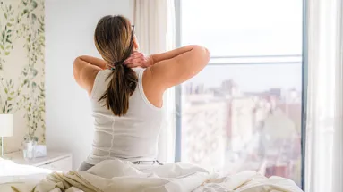 woman sitting on edge of bed with arms behind kneck looking out of the window