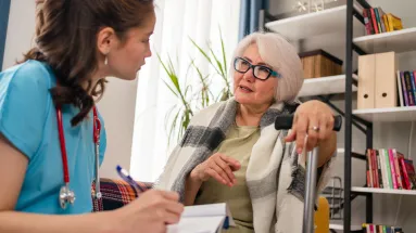 Nurse writing on note pad talking to older woman who is sitting down. 