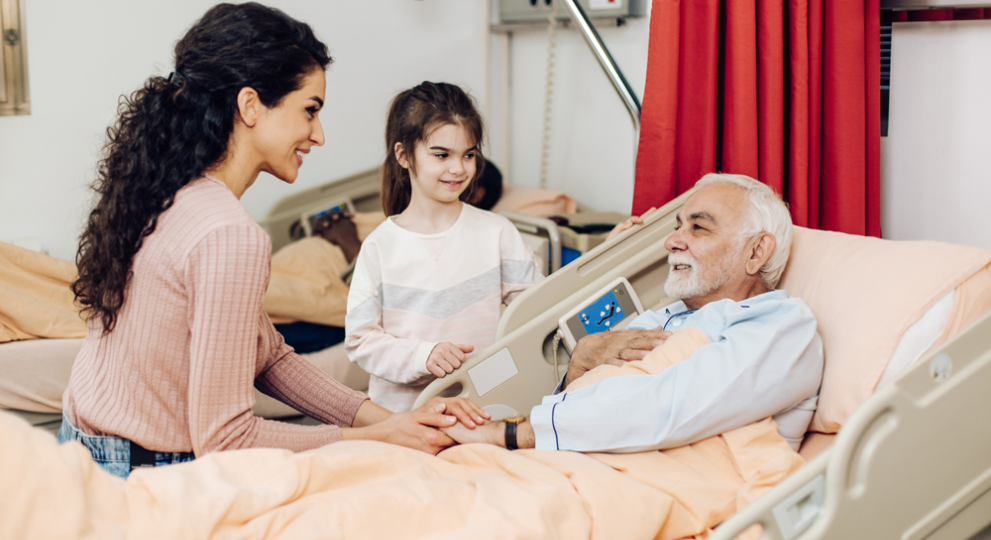 Man laying in hospital bed with a woman and female child by his bed side.