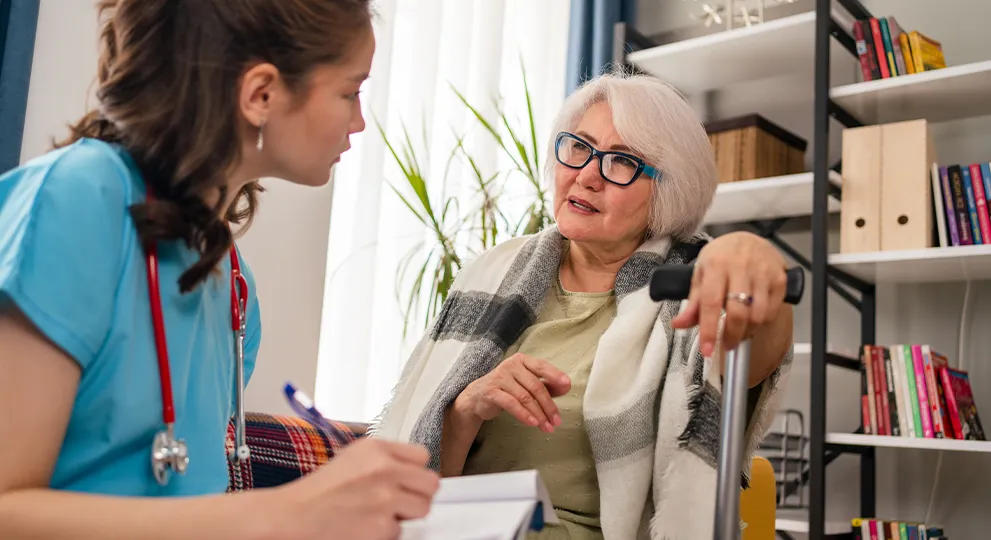 Nurse writing on note pad talking to older woman who is sitting down.